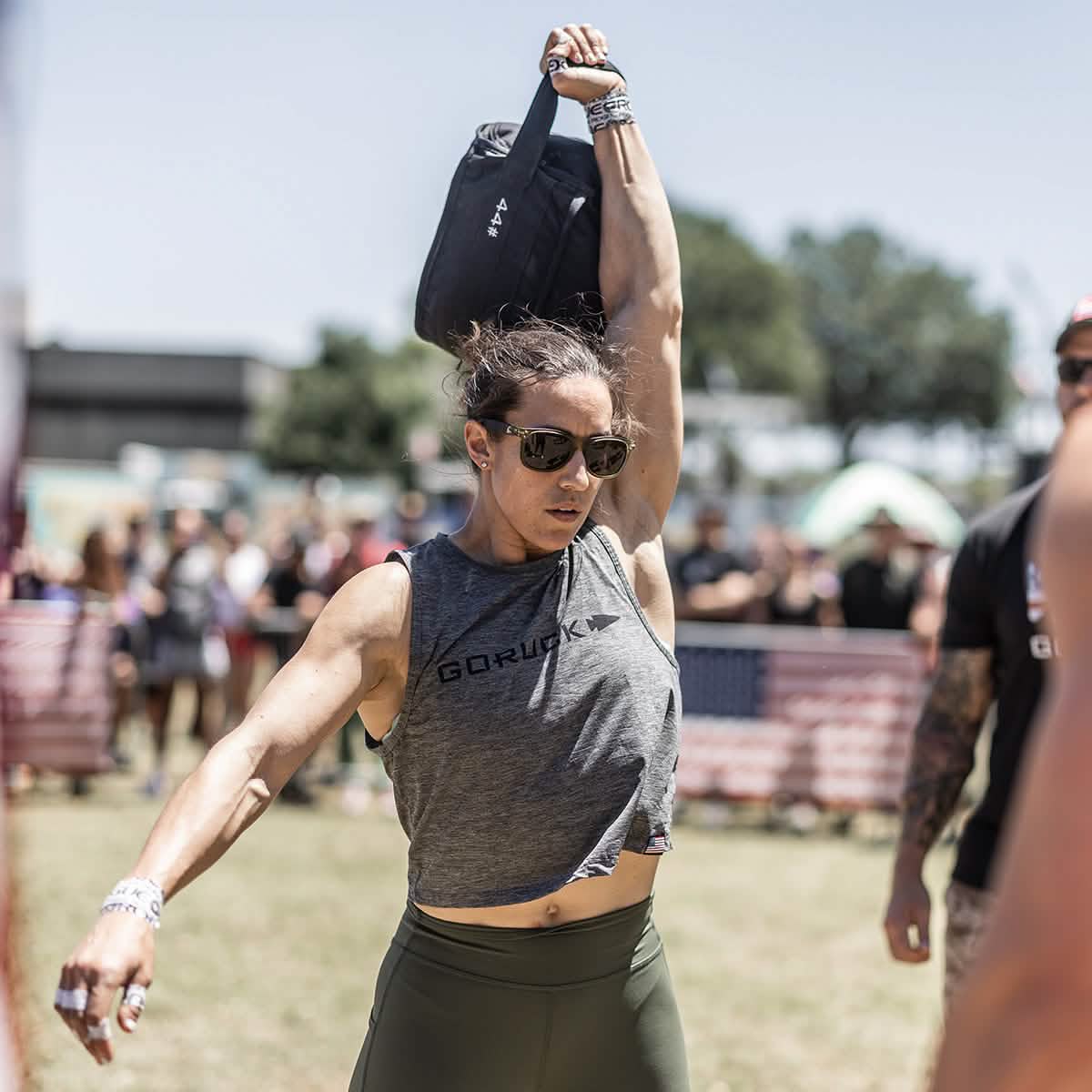 Athletic woman wearing sunglasses and gray GORUCK tank top lifting a black weighted bag outdoors at fitness event