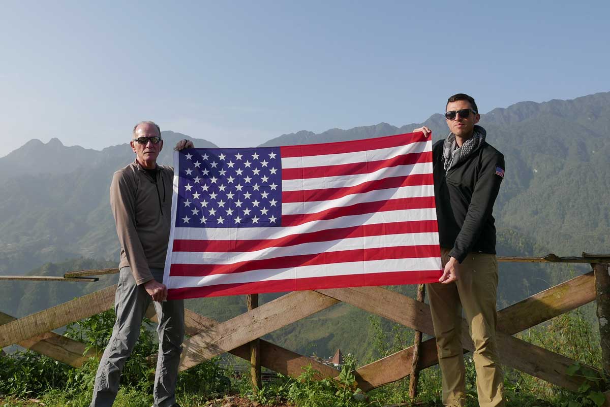 Two men holding a US flag outdoors with mountains in the background, GORUCK adventure gear.