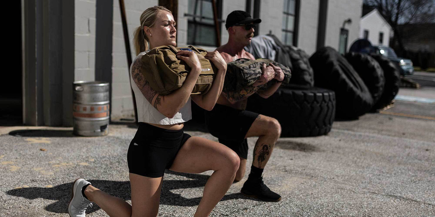 Two fit adults doing weighted lunges outside gym with sandbags near large tires and keg
