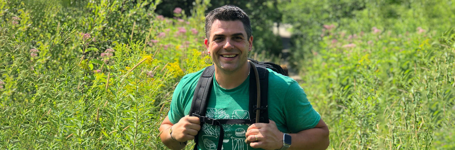 Man rucking outdoors in GORUCK gear, smiling on a green trail surrounded by wildflowers