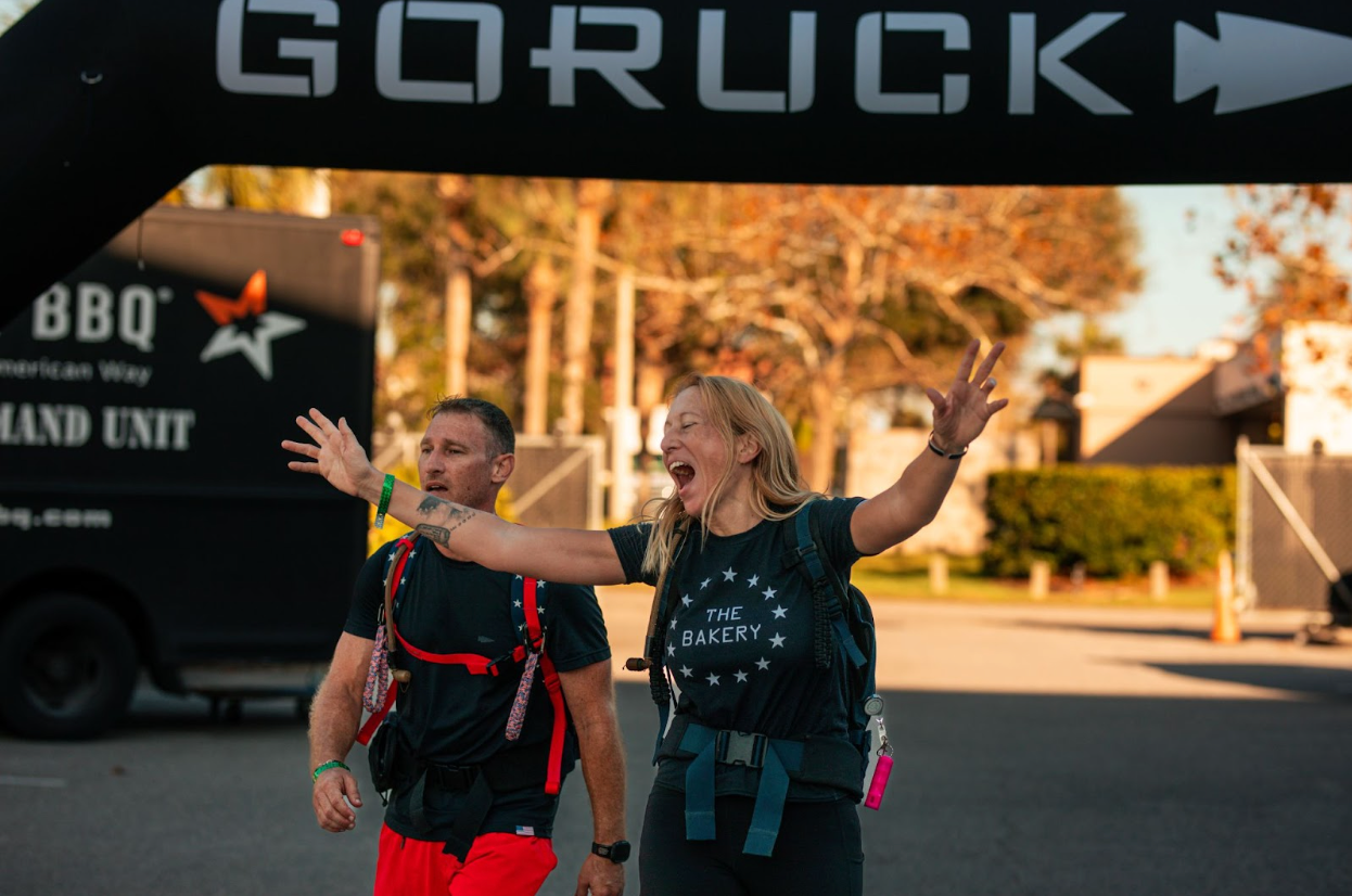Two adults with backpacks under a GORUCK event arch, woman cheering with arms wide open