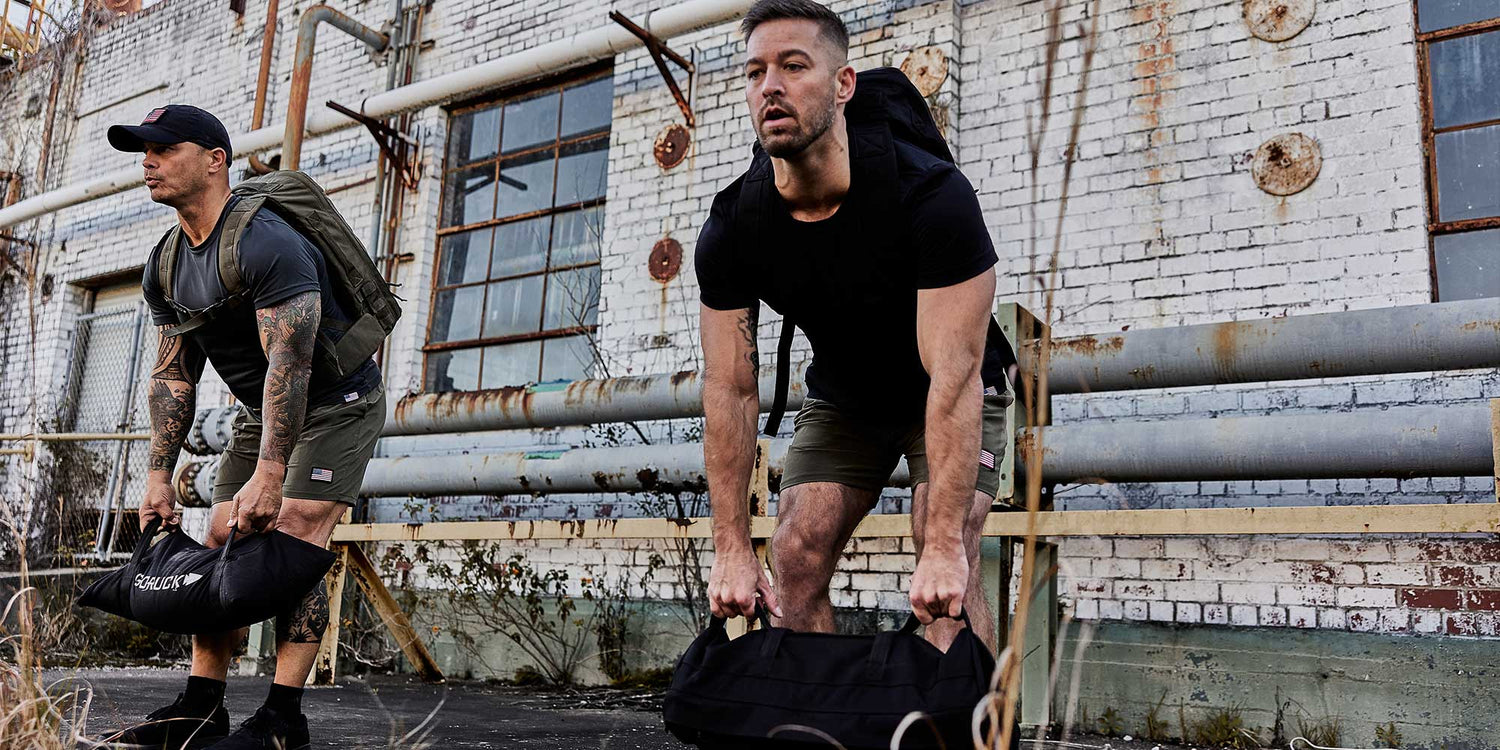 Two men rucking with GORUCK gear and sandbags in front of an industrial brick building