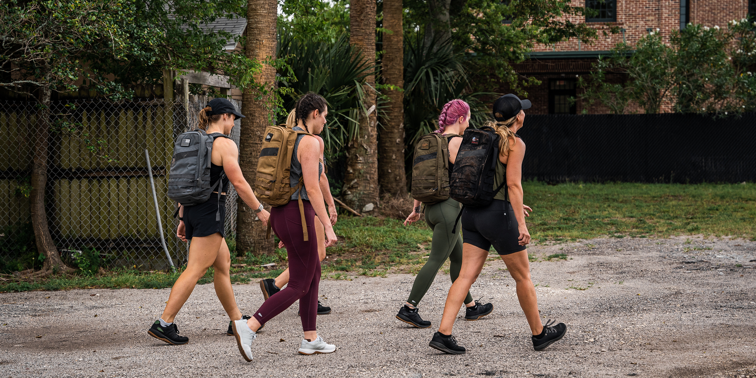 Four women walking outdoors wearing GORUCK backpacks and athletic gear in a green, urban park setting