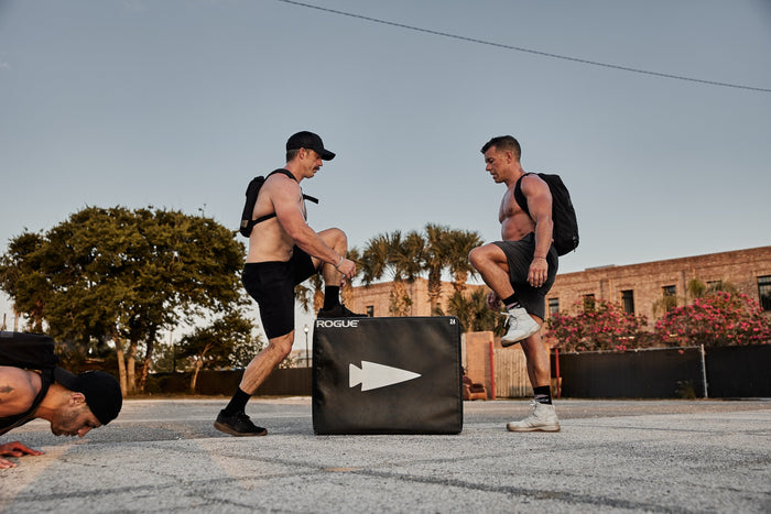 Two men wearing GORUCK rucksacks exercise outdoors with a Rogue plyo box, another man doing push-ups.