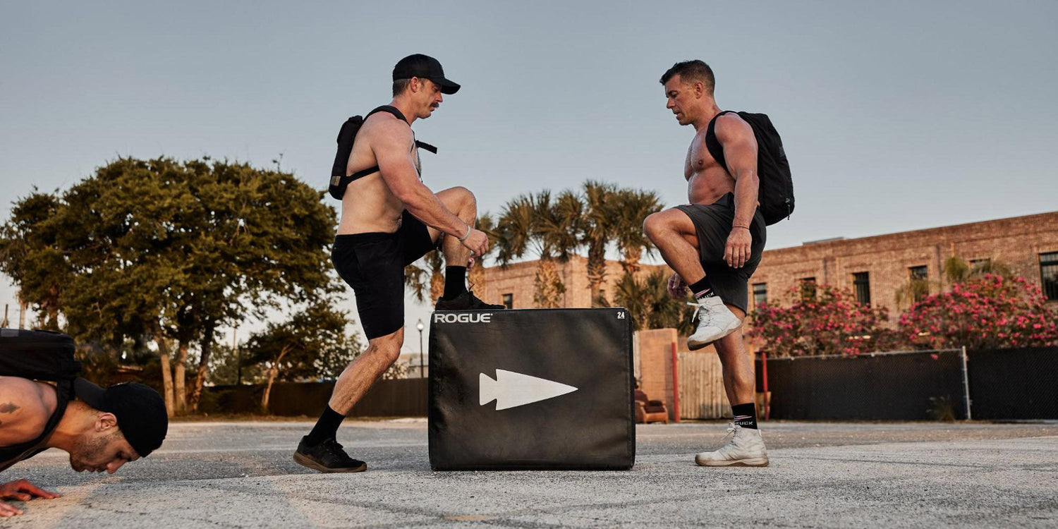 Two shirtless men wearing backpacks and athletic shorts warming up with box step exercises outdoors on a paved area