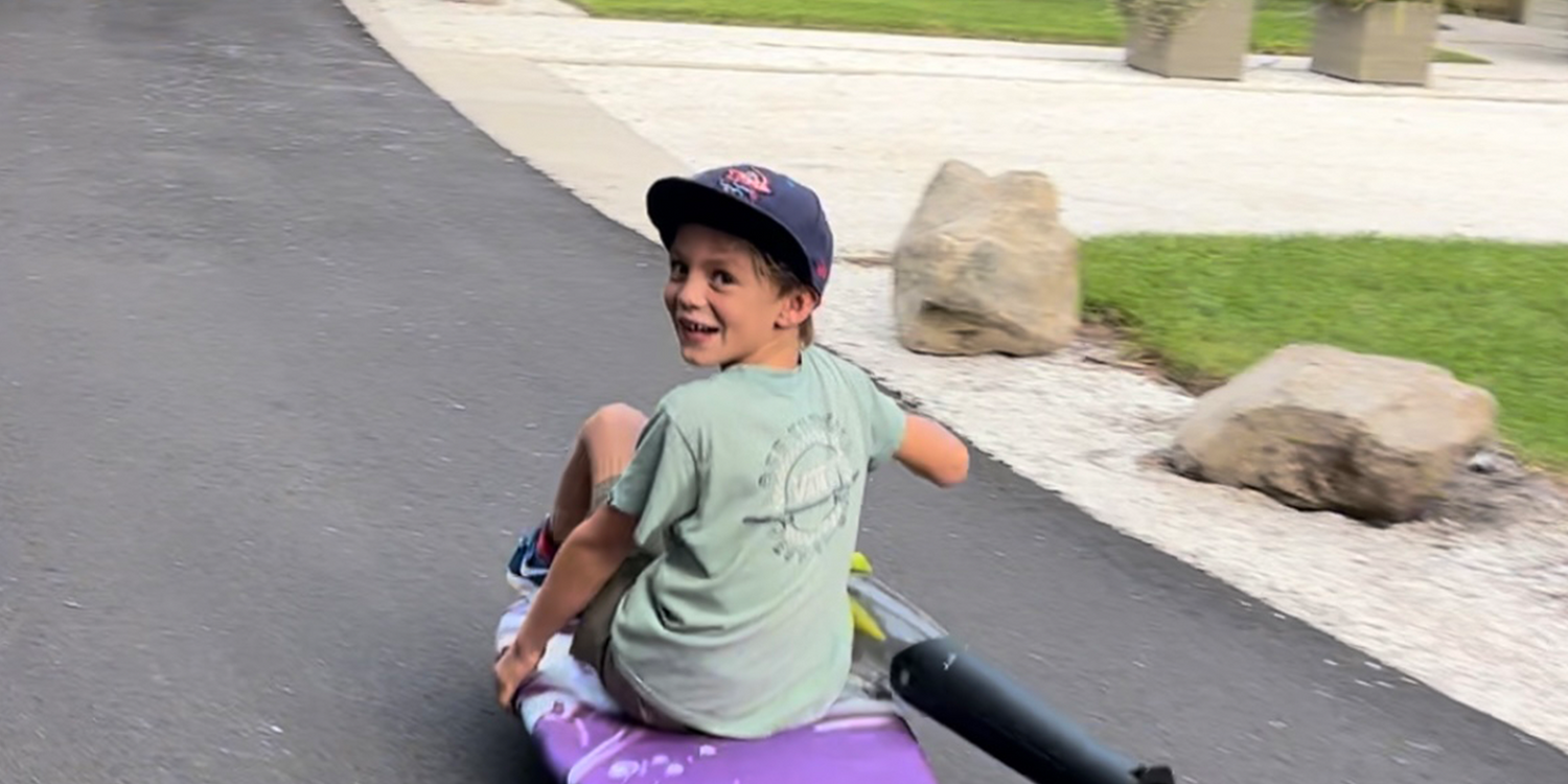 Young boy in cap riding a homemade scooter board on a paved path outdoors