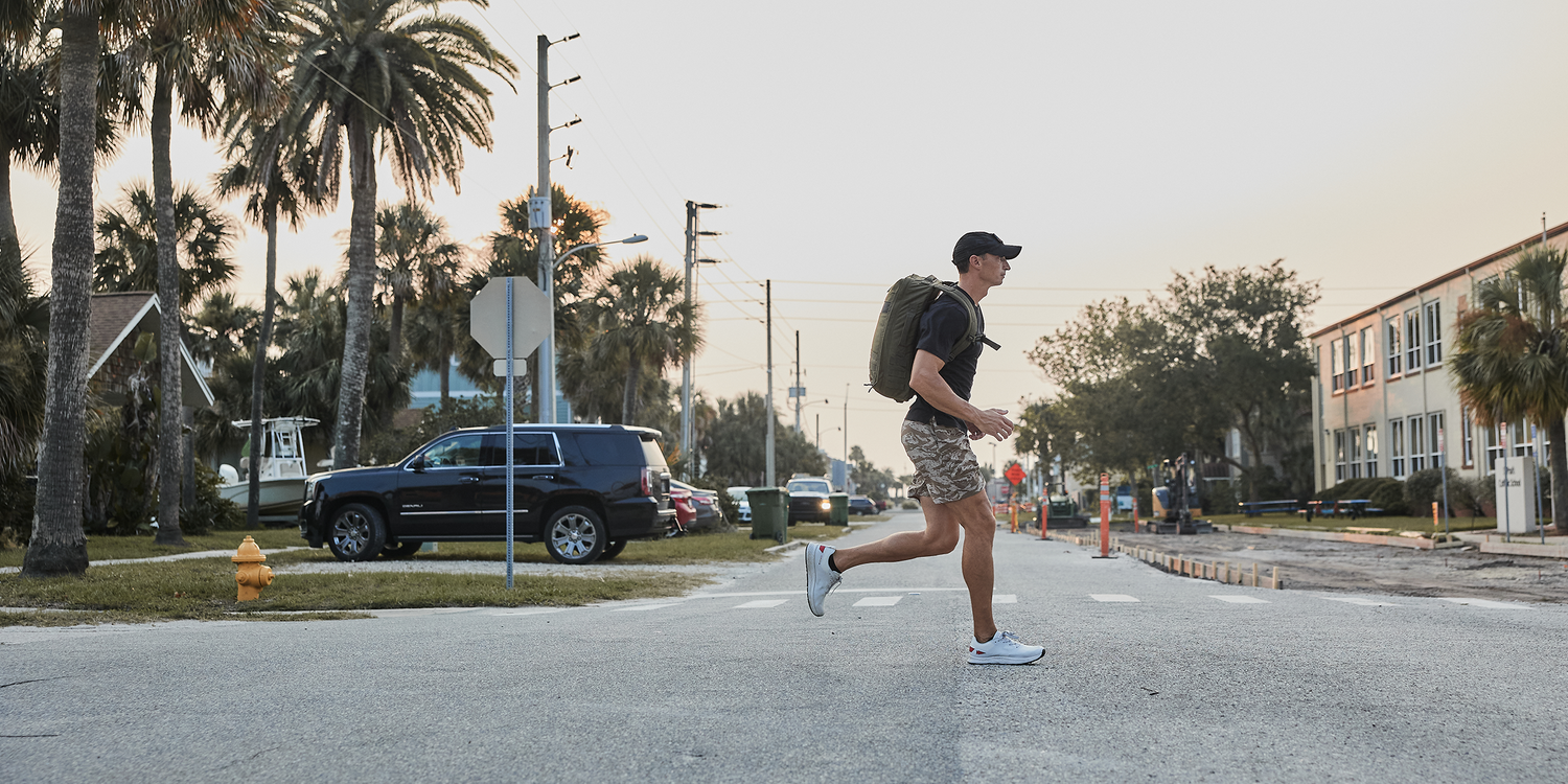 Man rucking with GORUCK backpack and camo shorts crossing urban street lined with palm trees