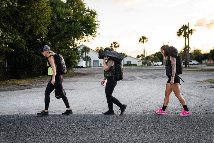 Three women rucking with GORUCK gear outdoors at sunrise, carrying weighted backpacks and sandbags.