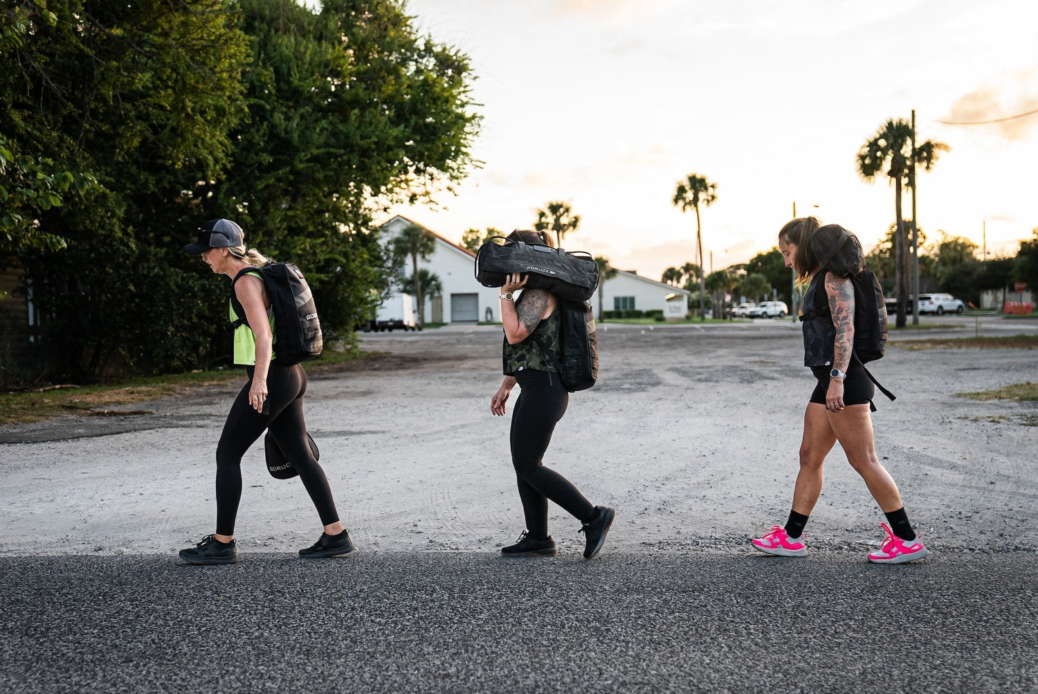 Three women walking outdoors carrying black weighted backpacks for fitness training at sunset