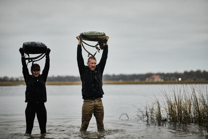 Two people rucking in water, holding GORUCK backpacks overhead outdoors