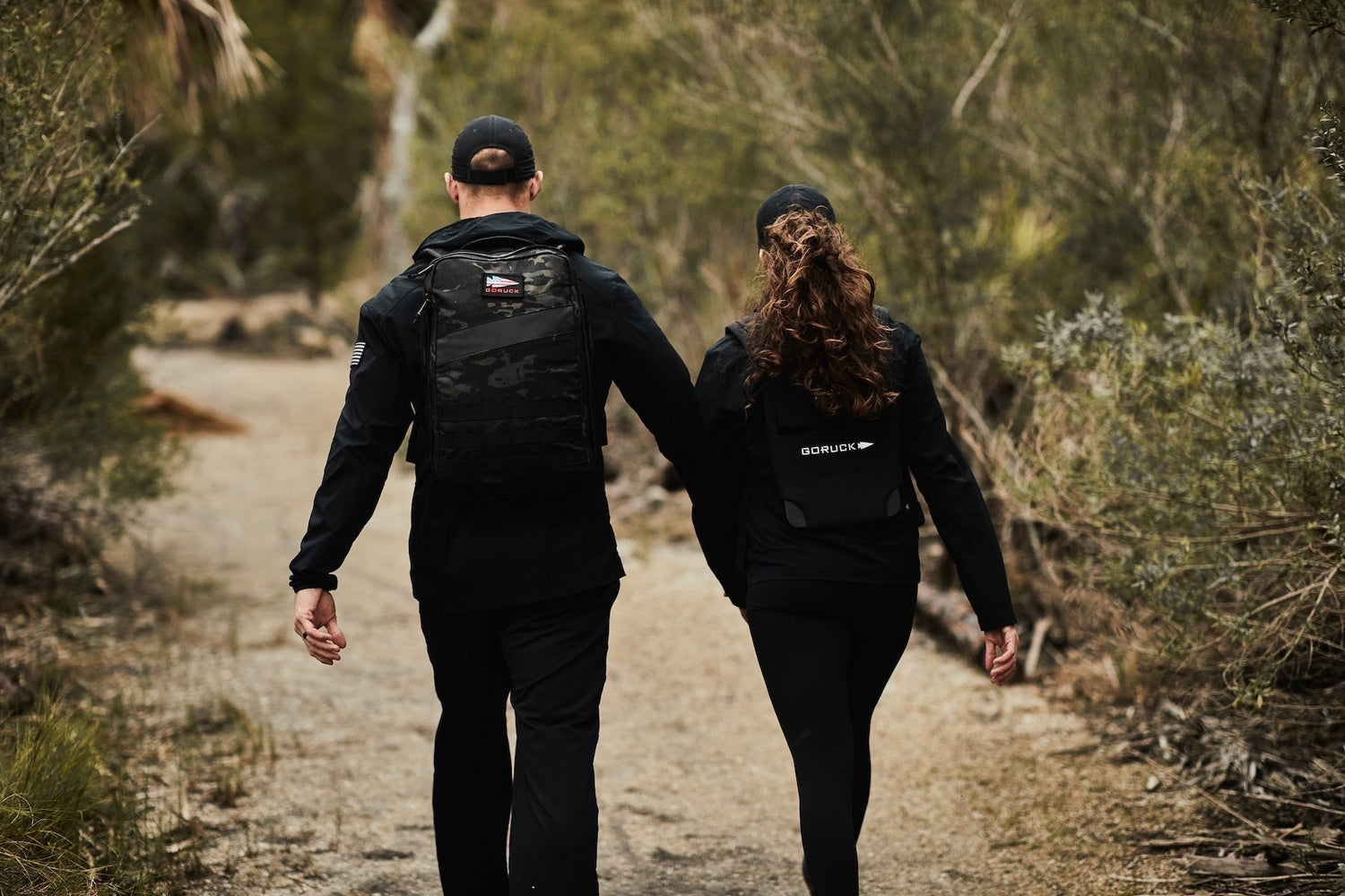 Two people wearing GORUCK backpacks walking on a trail surrounded by greenery