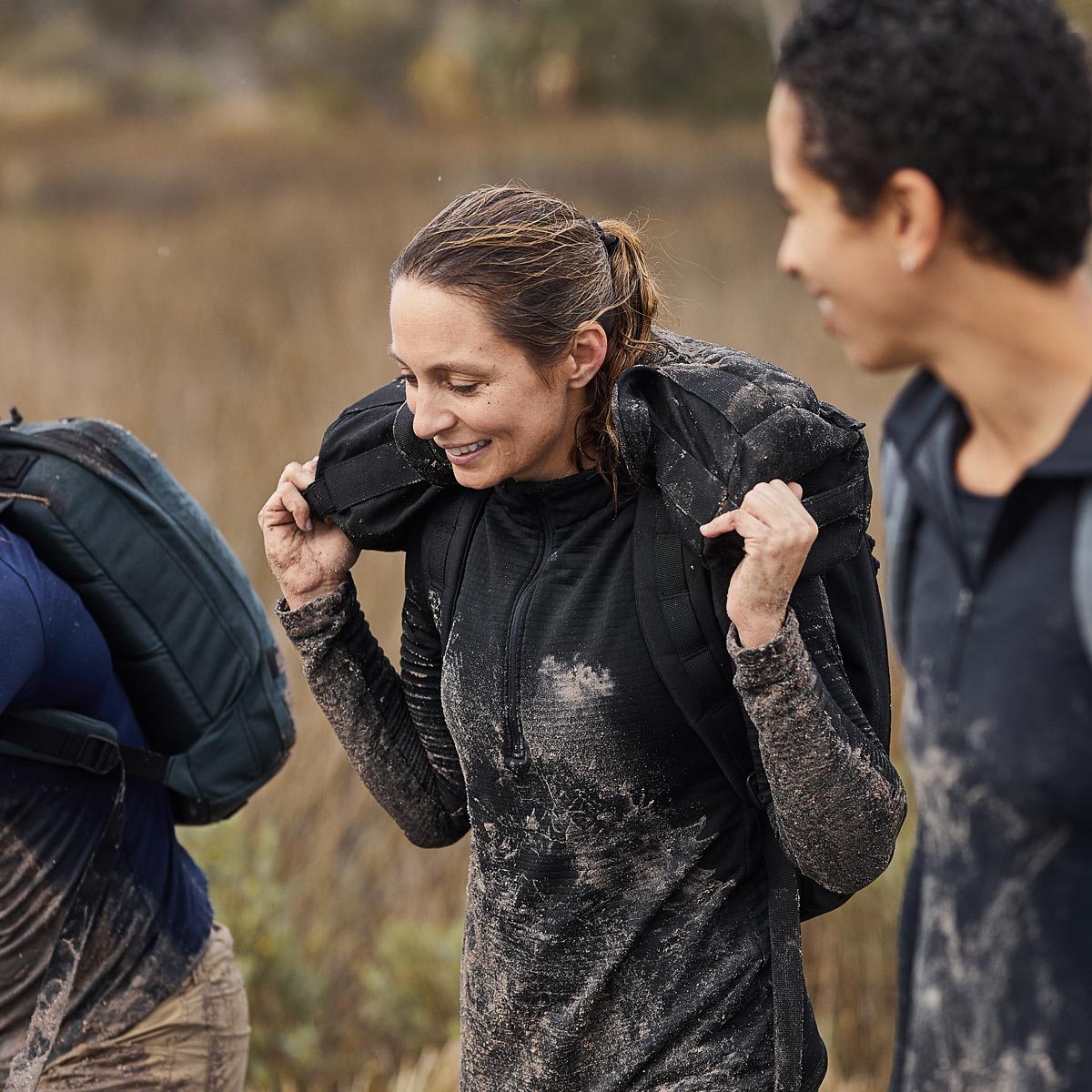Smiling woman in black mud-stained activewear carrying weighted sandbag outdoors, with two others nearby