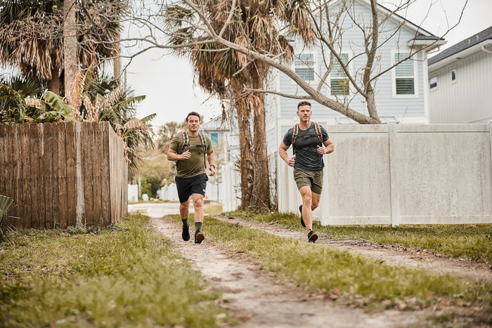 Two men rucking outdoors with GORUCK backpacks on a dirt path by palm trees and houses