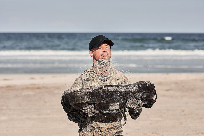 Man lifting GORUCK sandbag on sandy beach, wearing cap and rucking gear near ocean