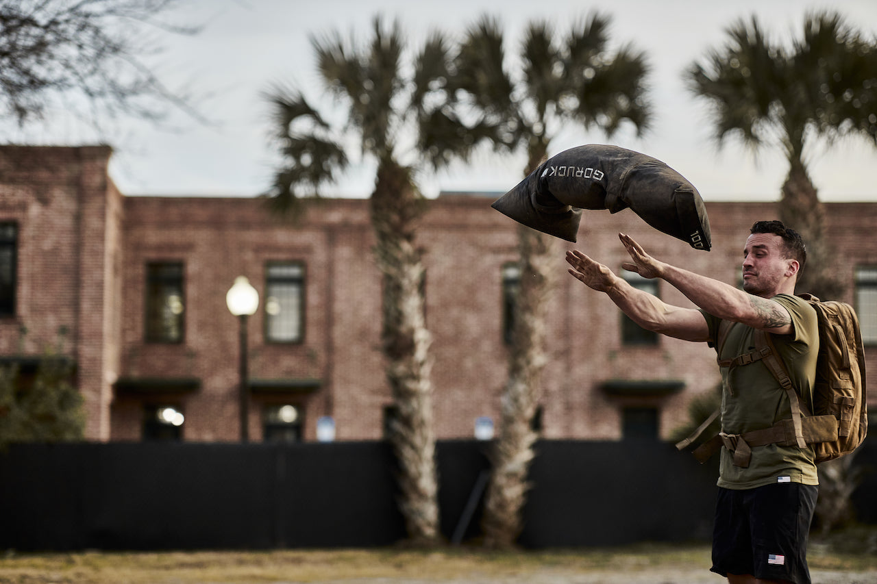 Man outdoors tossing GORUCK sandbag, wearing backpack, palm trees and brick building in background