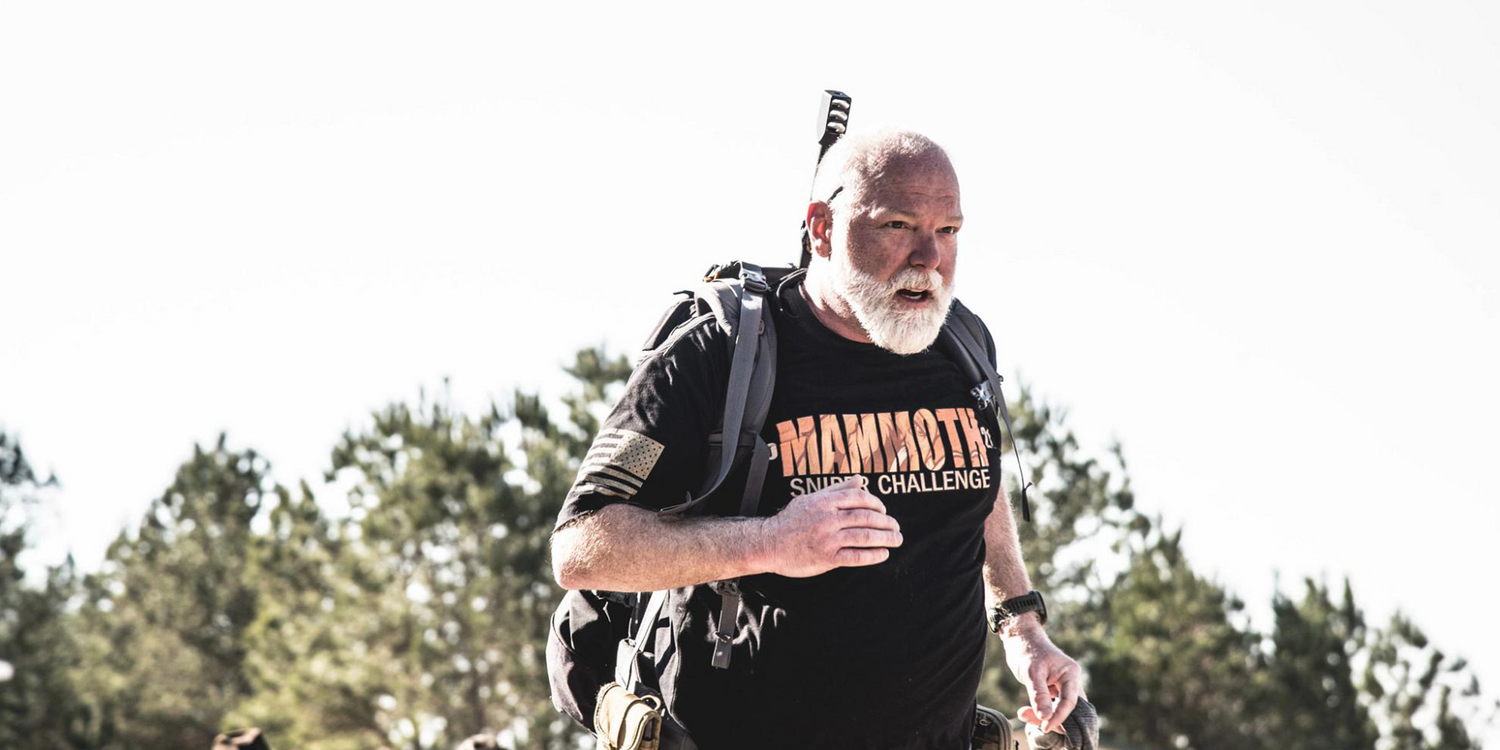 Man rucking outdoors wearing GORUCK gear and Mammoth Sniper Challenge shirt, trees in background