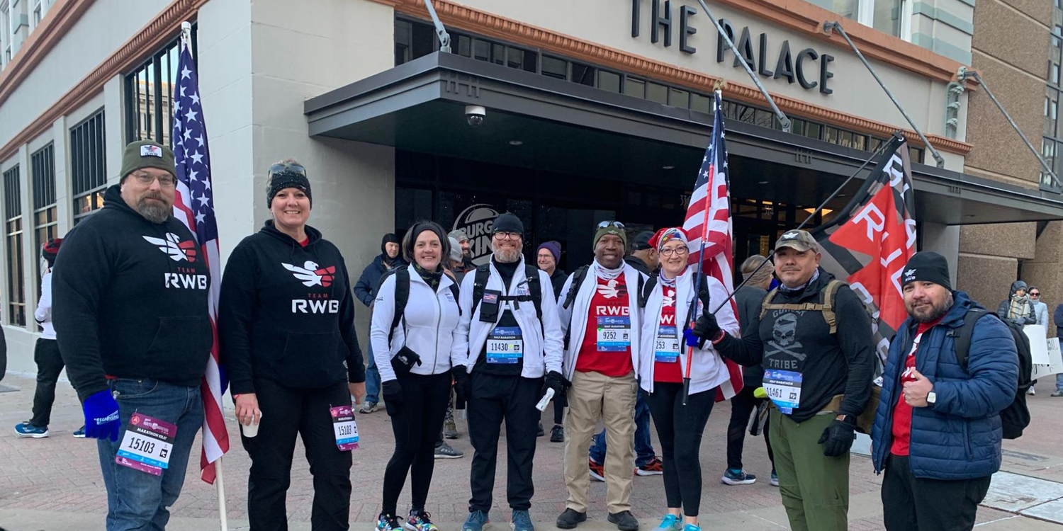 Group of diverse marathon runners wearing Team RWB gear with American flags outside The Palace building