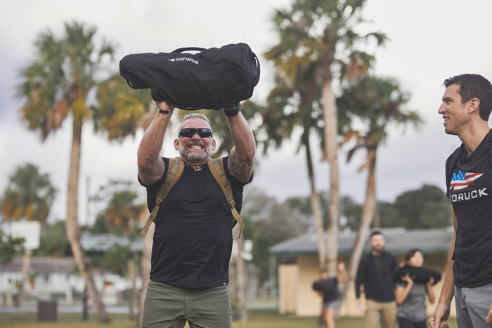 Smiling man lifts GORUCK sandbag outdoors during group rucking training with palm trees