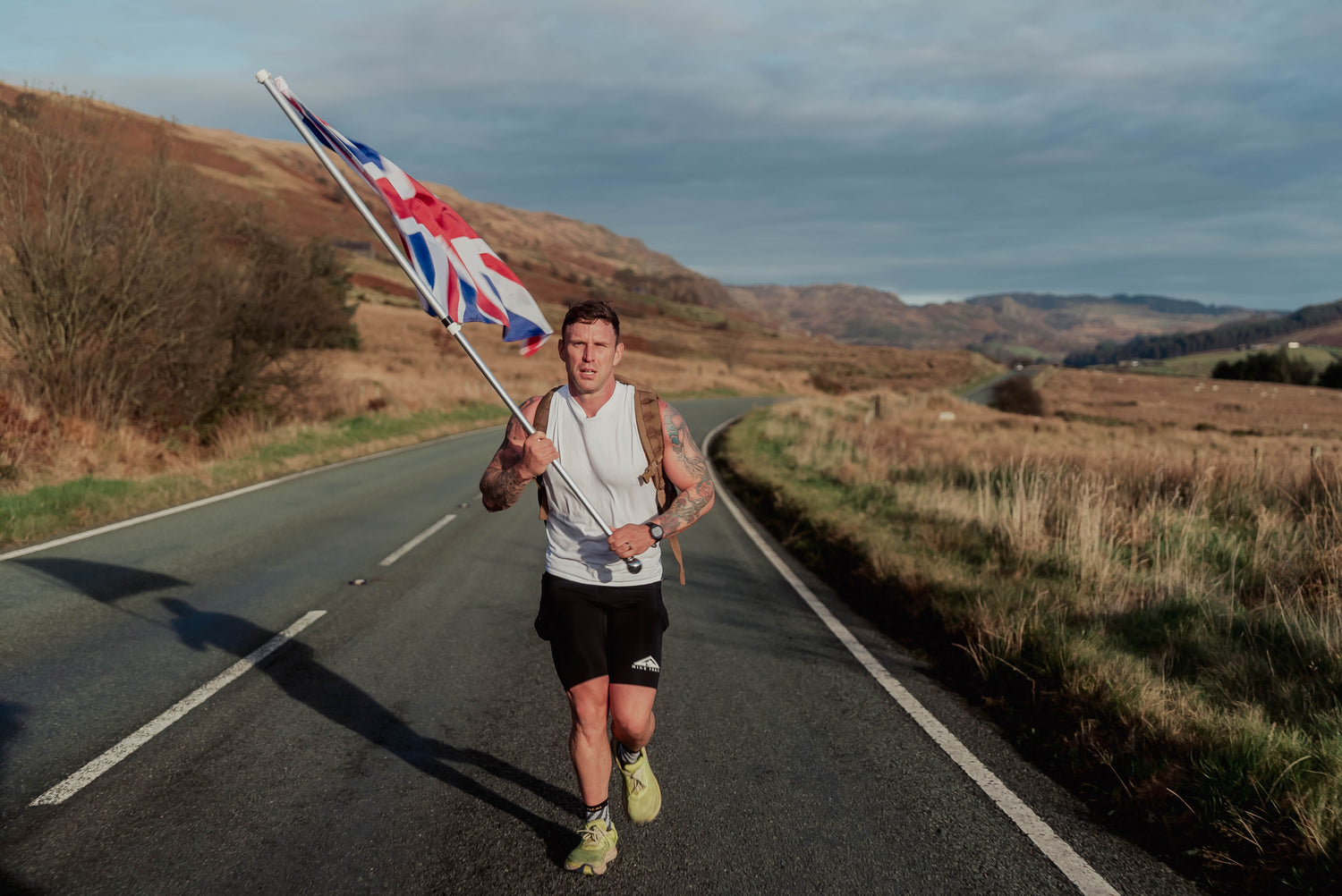 Man rucking on rural road carrying Union Jack flag, wearing GORUCK gear and athletic wear