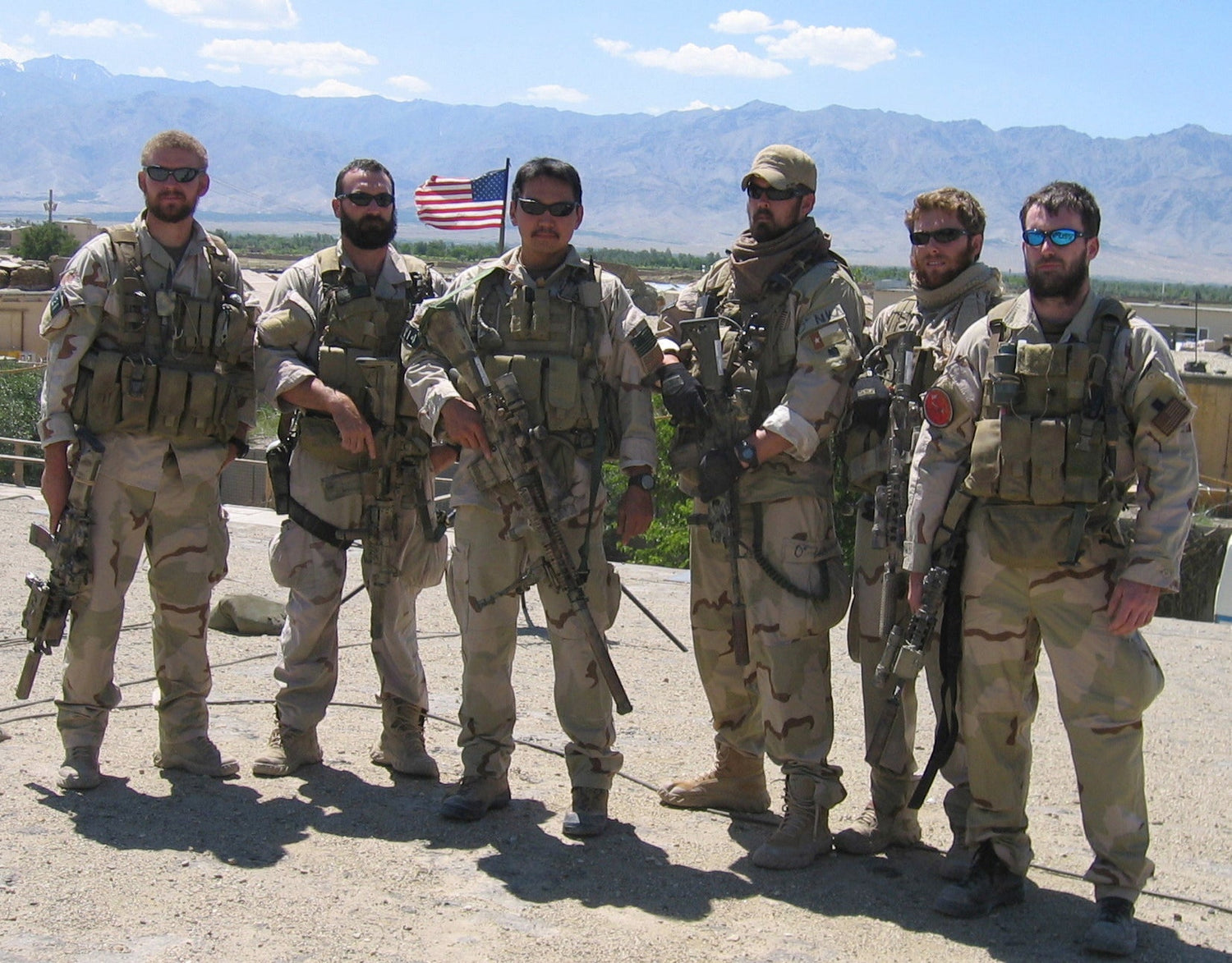 Six men in tactical gear and camouflage stand outdoors, mountains and US flag in background, GORUCK