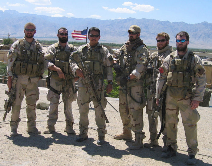 Six men in tactical gear and camouflage stand outdoors, mountains and US flag in background, GORUCK