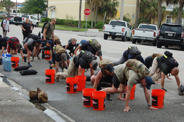 Group of people wearing GORUCK backpacks doing crawling exercises outdoors next to orange GORUCK buckets on a street
