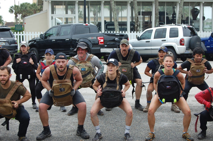 Group of people wearing rucking backpacks doing squats in outdoor parking area during GORUCK training event