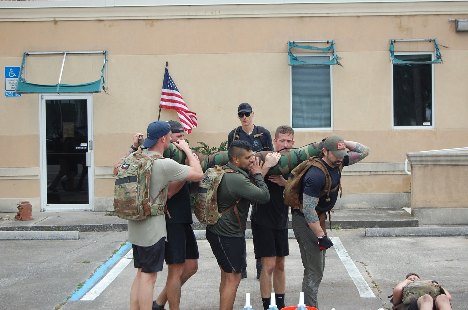 Group of men with GORUCK rucksacks lifting sandbag outdoors, American flag in background