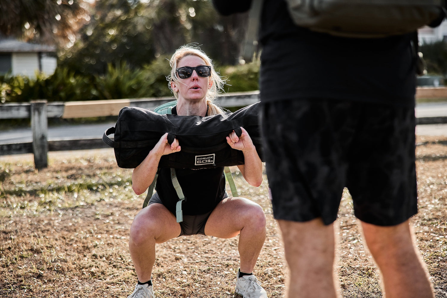 Woman doing outdoor rucking squat with GORUCK sandbag, wearing backpack and sunglasses