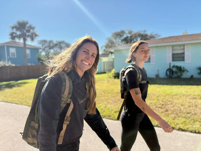 Two women rucking with GORUCK gear on a sunny suburban street, smiling and walking past homes
