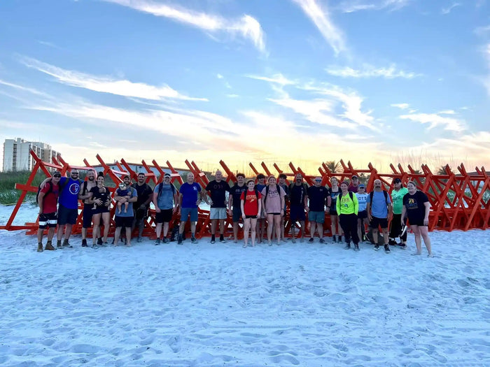 Group of people in casual and athletic gear standing on sandy beach near stacked red rucking barriers at sunset
