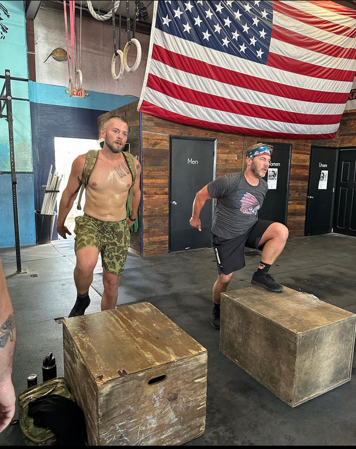 Two men doing box step exercises in a gym with wooden plyo boxes under a large American flag