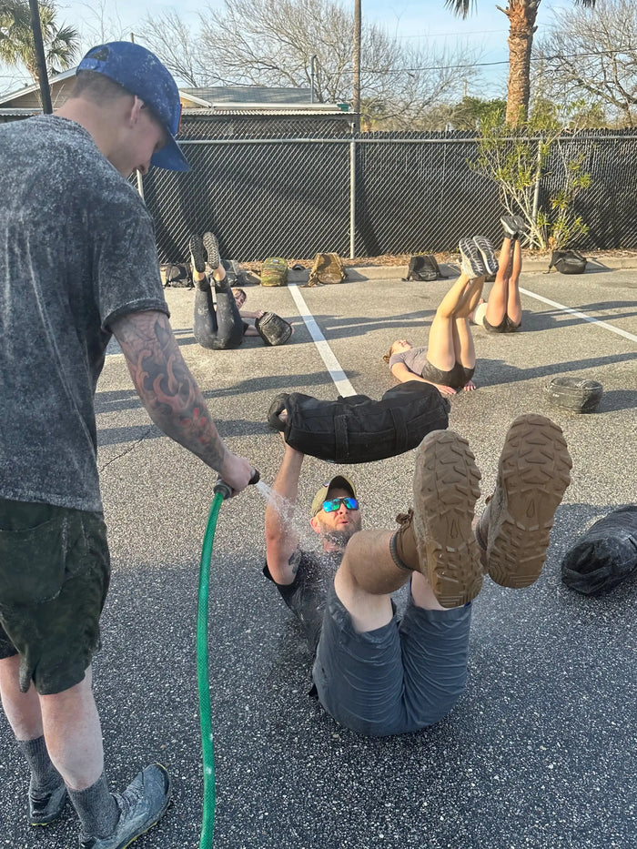 Group rucking workout in parking lot with sandbags and water spray, showing outdoor fitness training