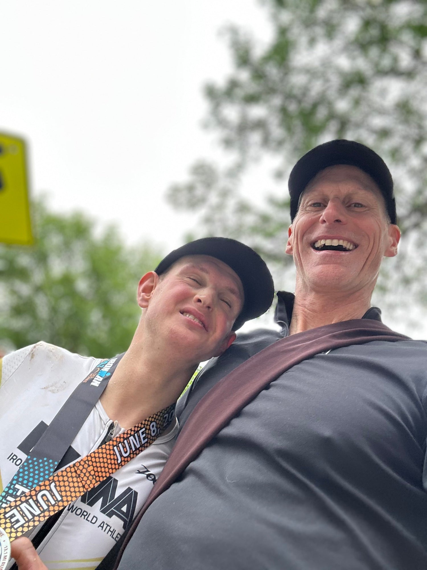 Two smiling men wearing hats, one with a race medal, outdoors under trees, GORUCK community spirit