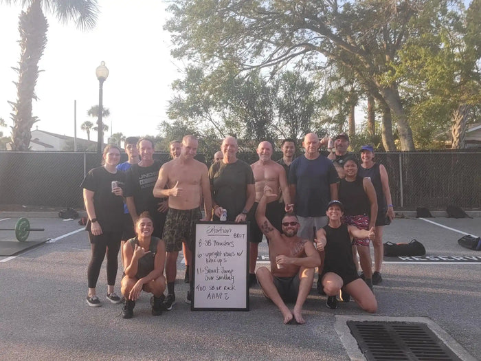 GORUCK group of men and women posing outside after a fitness training event, with a whiteboard workout list.