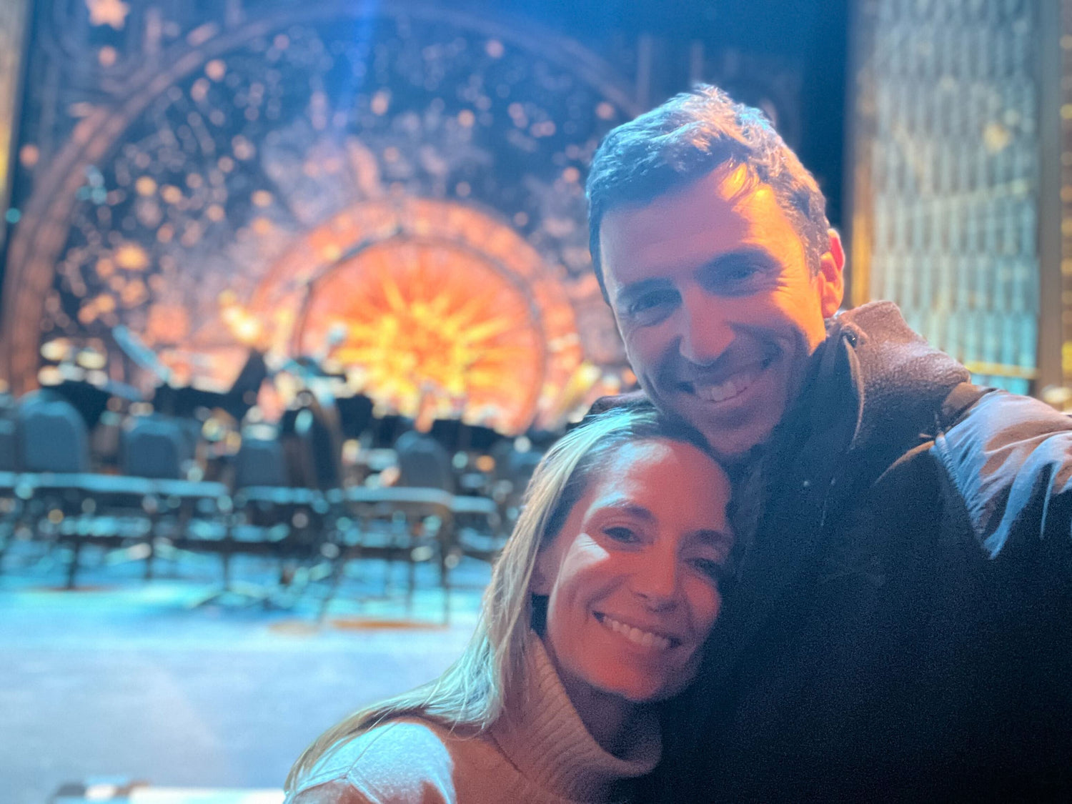 Smiling couple taking a selfie indoors with blurred stage featuring sunburst light and chairs in background