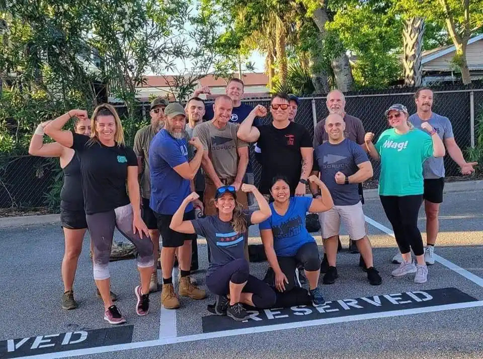 Group of diverse men and women flexing muscles outdoors in a parking lot with trees and fence background