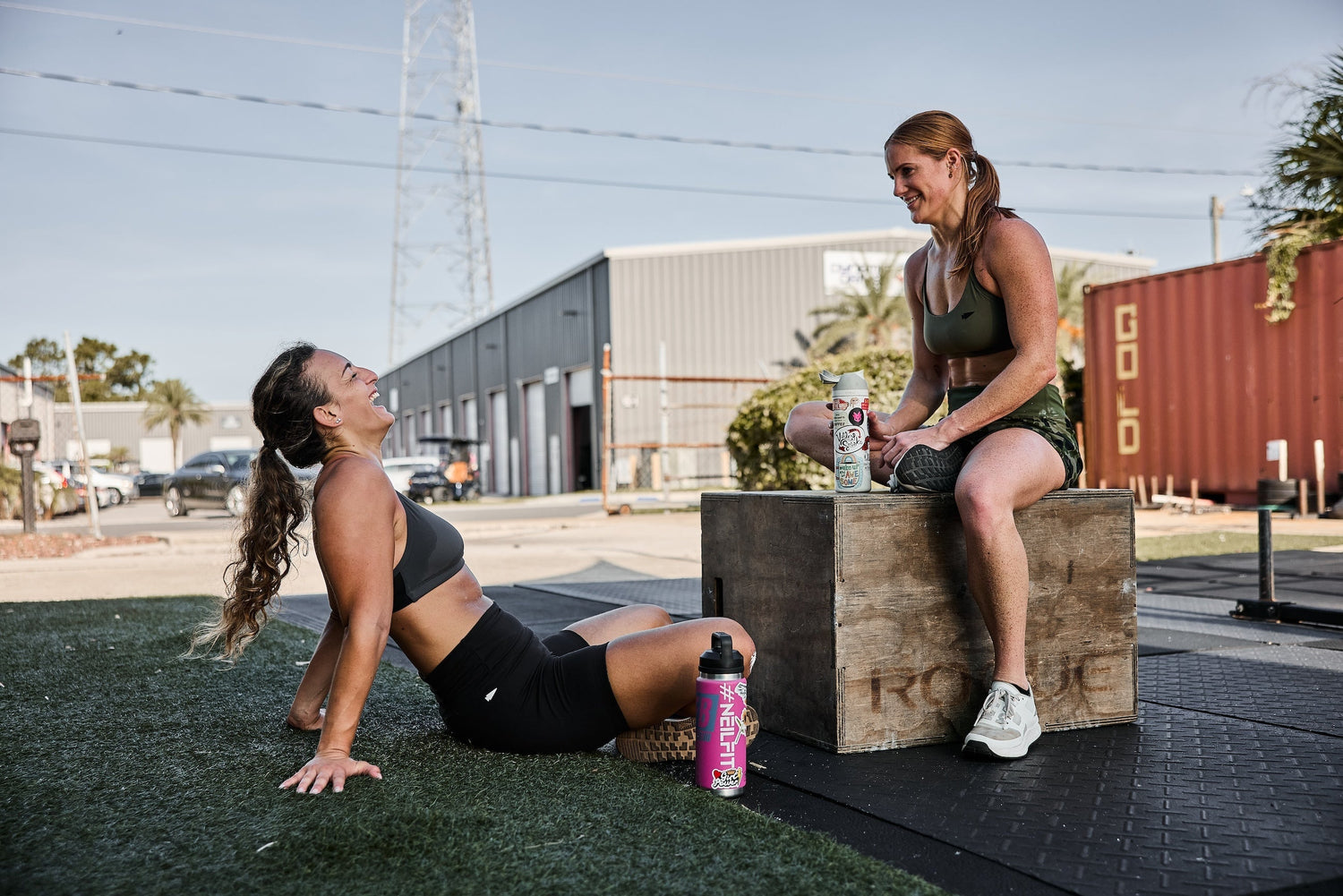 Two fit women in sportswear relaxing and laughing outdoors near gym equipment and water bottles