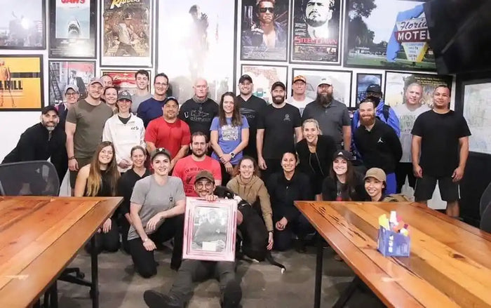 GORUCK team group photo in office with movie posters on wall, wooden tables, community spirit