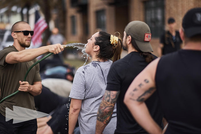 Outdoor GORUCK rucking training, woman drinks from hose, group in athletic gear.