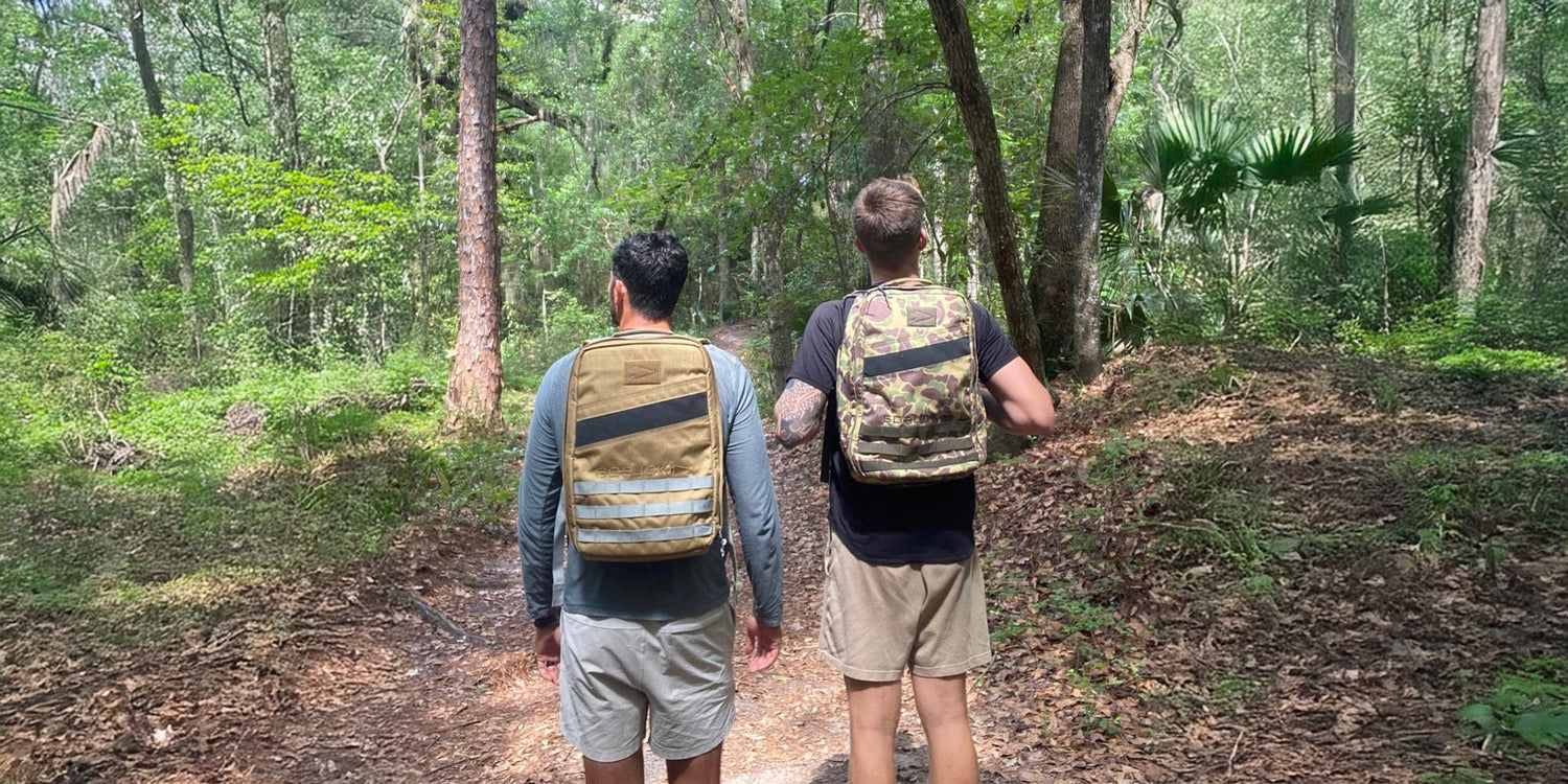 Two men with backpacks hiking on a forest trail surrounded by green trees and foliage