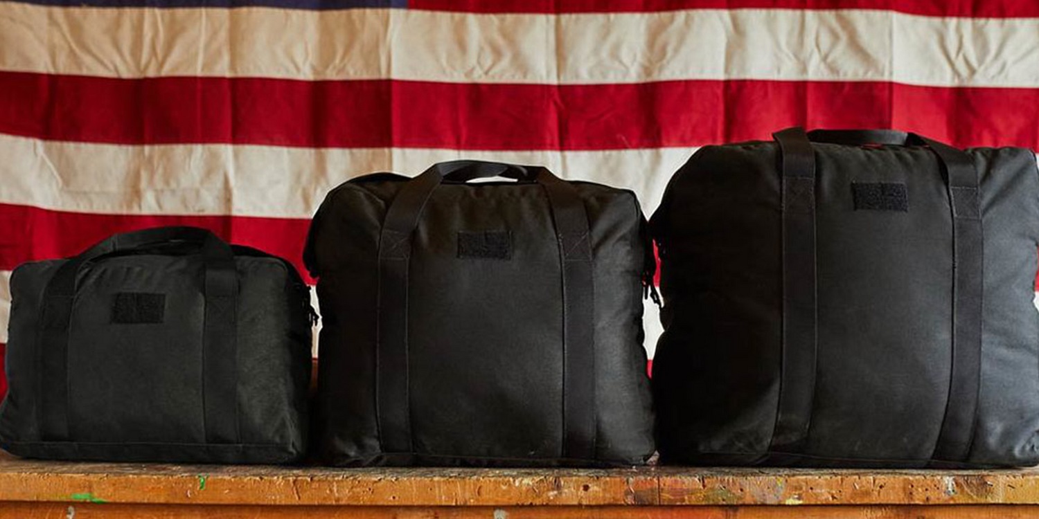 Three GORUCK black kit bags on a wooden table with an American flag backdrop.