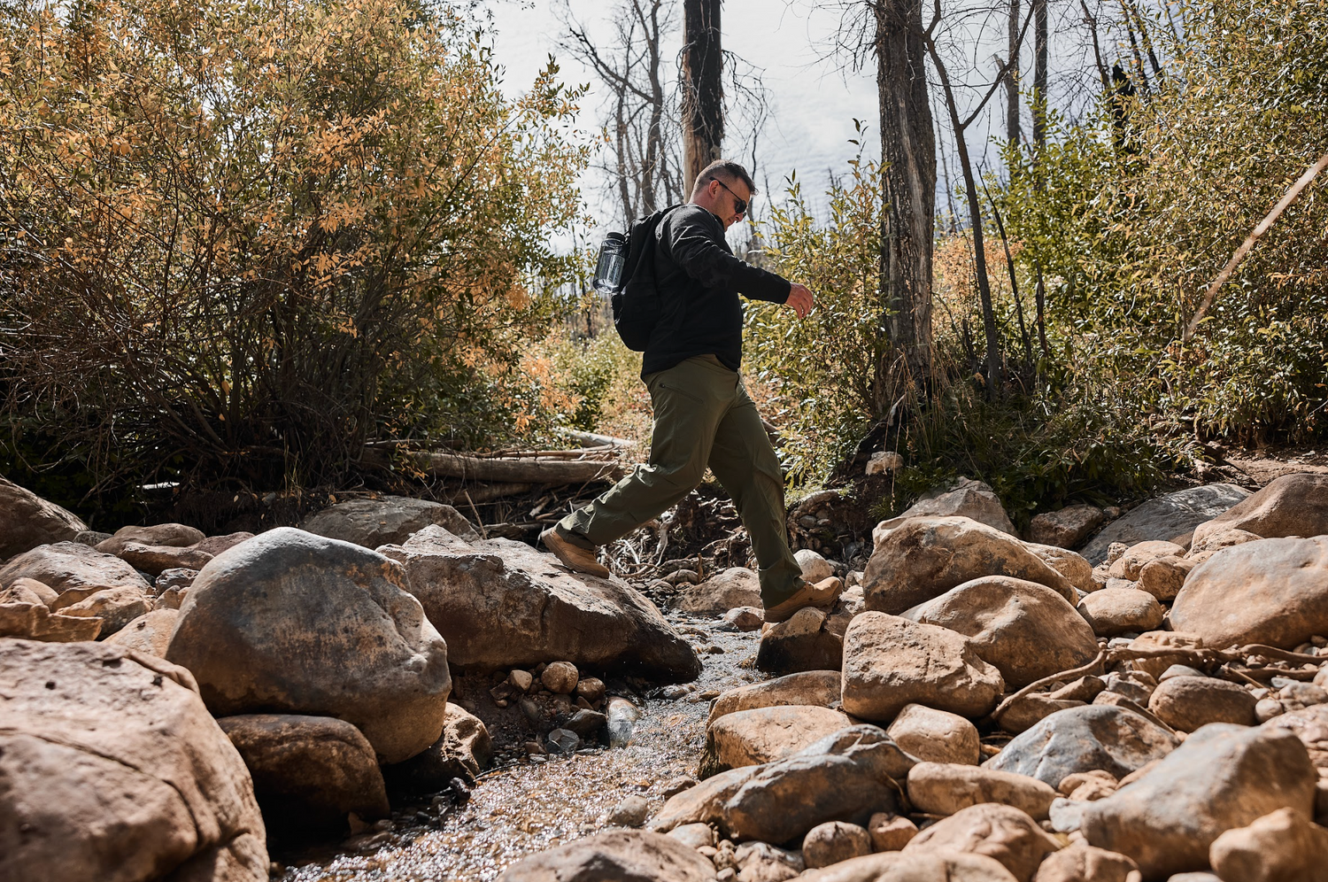 Man with backpack crossing rocky stream in autumn forest rucking outdoors