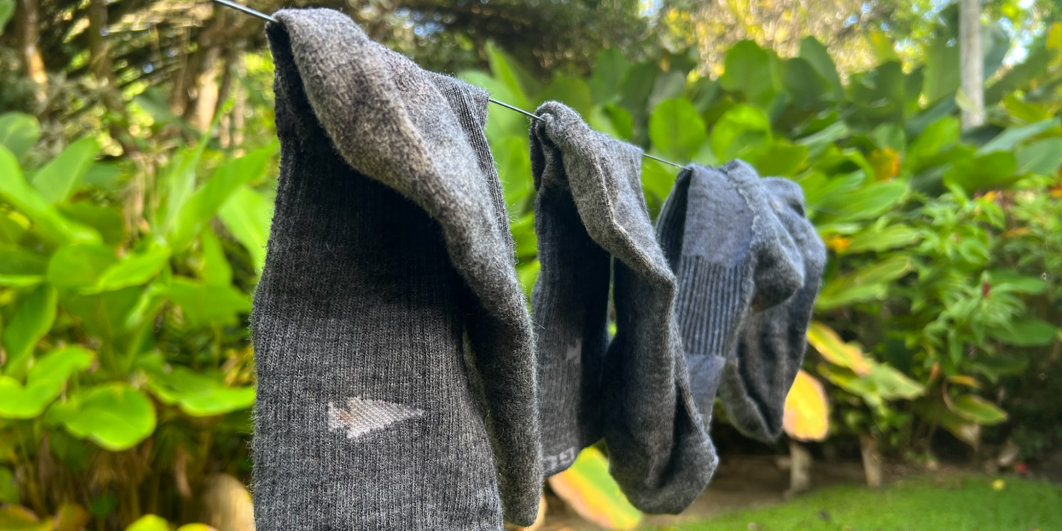 Three pairs of grey merino wool socks hanging on a clothesline outdoors with green foliage background