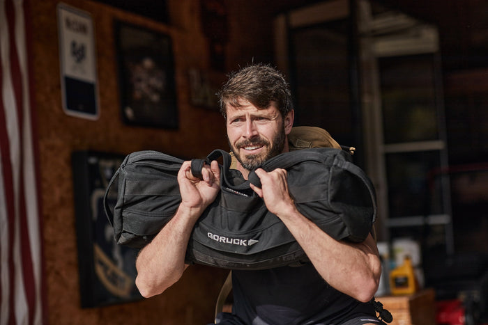 Man lifting GORUCK sandbag in garage gym, showcasing rugged rucking gear and training strength