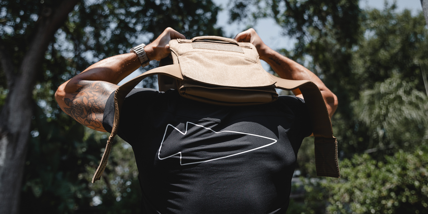 Person lifting tan GORUCK rucksack outdoors, wearing black shirt with arrowhead logo