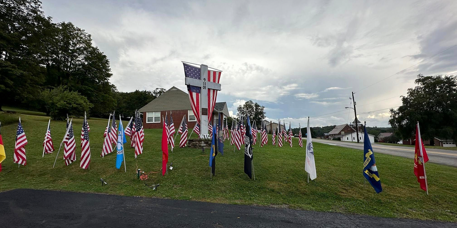 9/11 memorial with American flags and a cross on a grassy hill near a brick house under a cloudy sky
