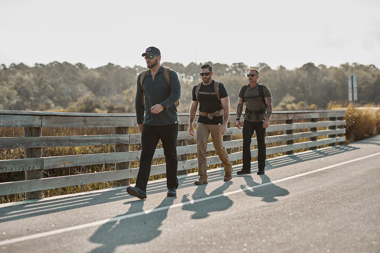 Three men rucking outdoors on a paved path with GORUCK backpacks, wooded area in background