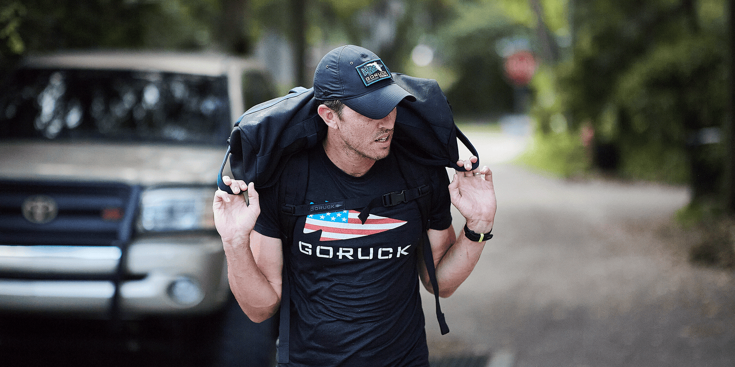 Man wearing GORUCK shirt and hat rucking with heavy backpack outdoors near parked vehicle