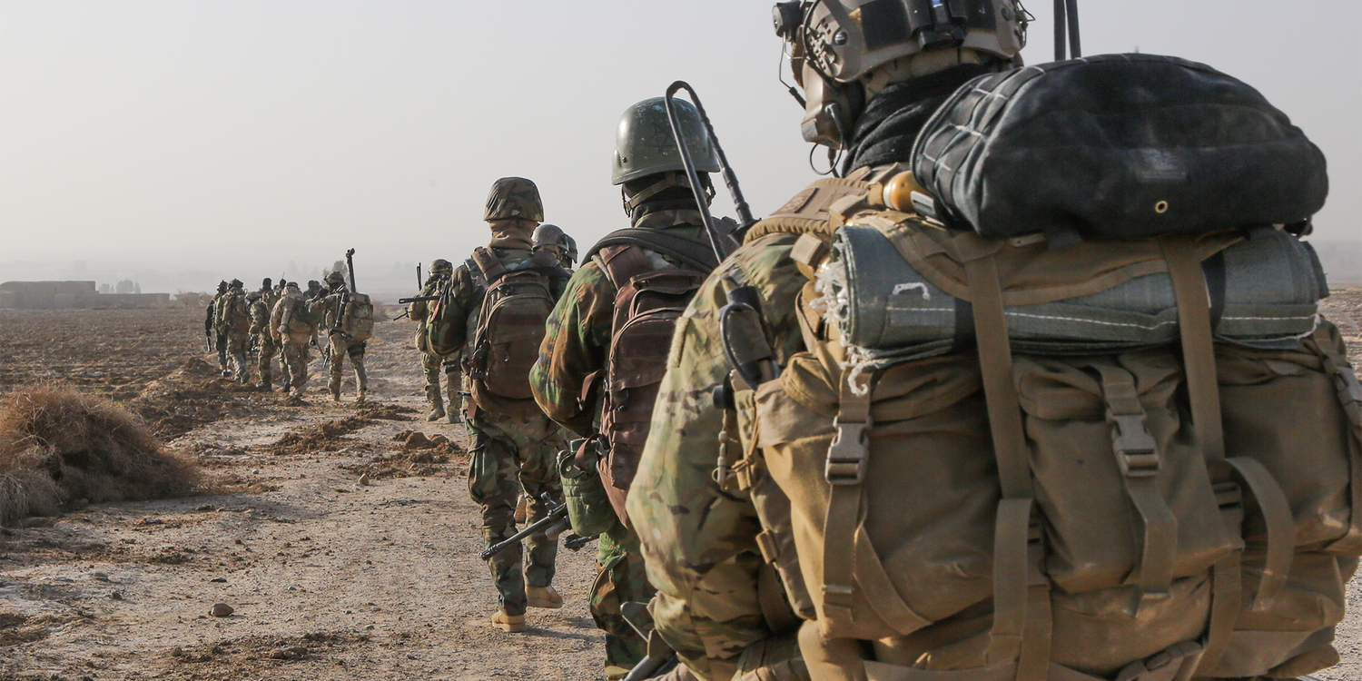 Group of soldiers in full military gear and backpacks rucking in a line across a barren field